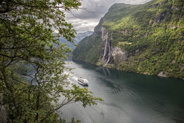 A cruise ship sails through the Gairanger Fjord surrounded by steep mountains and the Seven Sisters waterfall in Norway Europe