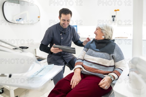 Dentist showing something on tablet to smiling senior woman sitting in dentist chair, during consultation in dental clinic