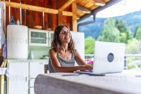 Happy freelancer woman working on laptop from the balcony of a mountain chalet, enjoying freedom and nature during summer vacation