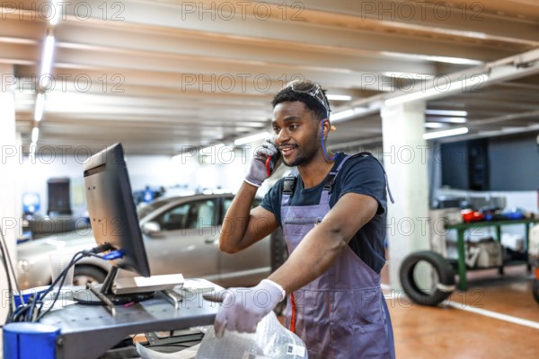 Young mechanic wearing gray uniform and protective glasses using computer and talking on phone while working in car service