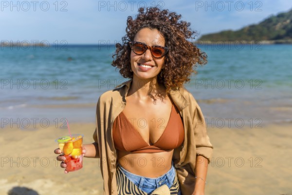 Confident arab woman holding a glass of tropical fruit at beach under the sun in a windy day