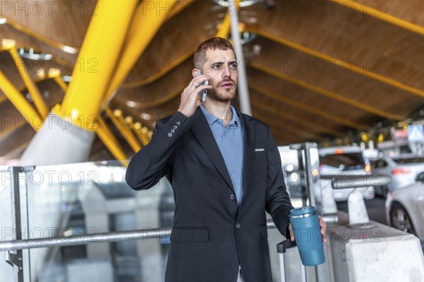Businessman in a suit holding a reusable coffee cup while using a smartphone, waiting at the airport for his next flight