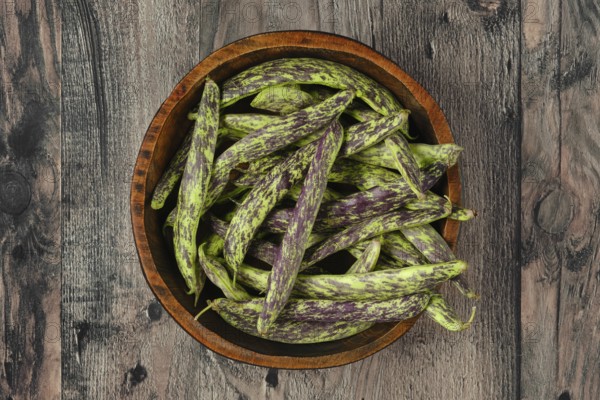 A wooden bowl filled to the brim with vibrant purple and green beans rests on a textured wooden surface. Dragon tongue beans in a bowl