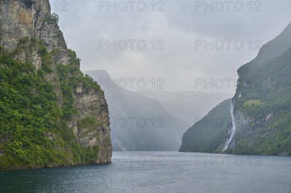 Geiranger Fjord with waterfall surrounded by rocks and mountains with overcast sky, Geiranger, Geiranger Fjord, Stranda, Romsdal, Norway, Europe