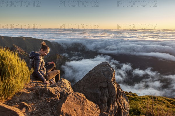 A woman sits on a rock and looks at the sea of clouds below her at dusk, Pico Ruivo, Madeira, Portugal, Europe