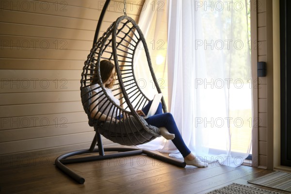 On a hot sunny day, a young woman sits in a hanging swing chair with a laptop in an air-conditioned room of log cabin