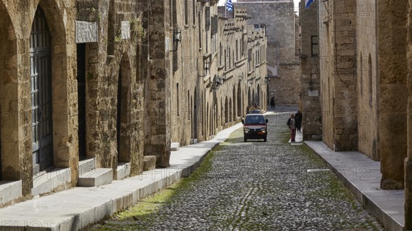 A historic cobblestone street with old yellow stone buildings, a moving car and a pedestrian, Knights Street, Rhodes Old Town, Rhodes Town, Rhodes, Dodecanese, Greek Islands, Greece, Europe