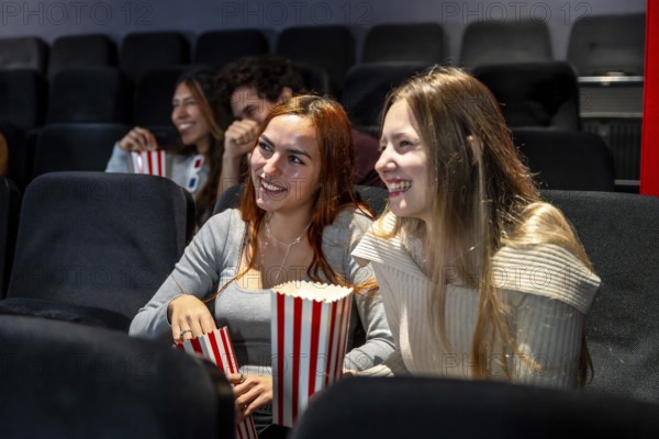 Two young women enjoying popcorn and smiling while watching a movie together in a cozy cinema, sharing laughter and a fun evening