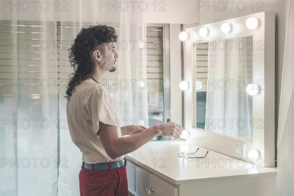 Young man with a mullet hairstyle standing in front of a lit mirror, holding items, dressed in a white t-shirt and red pants, preparing for the day in a cozy bedroom
