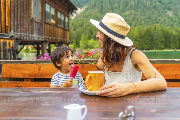 Mother and son enjoying a lakeside snack with a colorful popsicle and warm beverage against a stunning mountain backdrop