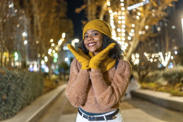 Smiling young woman wearing yellow woolen hat and gloves enjoying christmas lights in a city at night