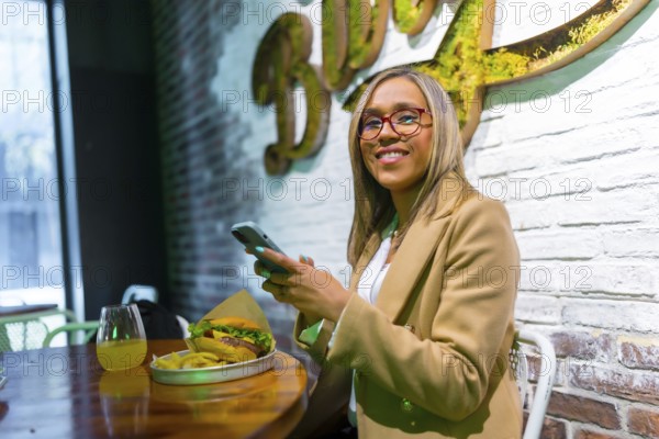 Latin woman smiling at camera sitting on a restaurant of fast food eating cheeseburger while using mobile phone