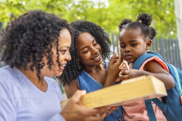 Cute african girl holding toys in arms of her mother next to her grandmother in a park