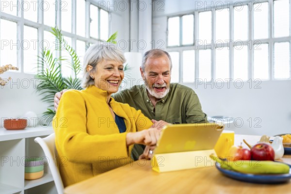 Happy senior couple using digital tablet at home pointing at the screen and smiling while eating breakfast together
