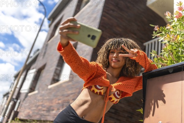 Latin student taking selfie gesturing success outside the house leaning in the entrance under the sun