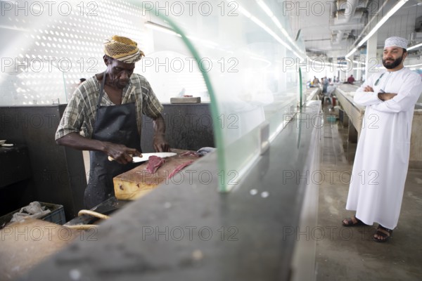 Omani man filleting a fish in Muttrah fish market, an Omani man in dishdasha watches, Muscat, Oman, Asia