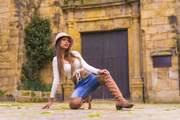 A young Latin woman with a white t-shirt, jeans and a hat enjoying the summer holidays, with a take away cafe, posado de moda, Pasaje San Juan, Gipuzkoa, Pais Vasco