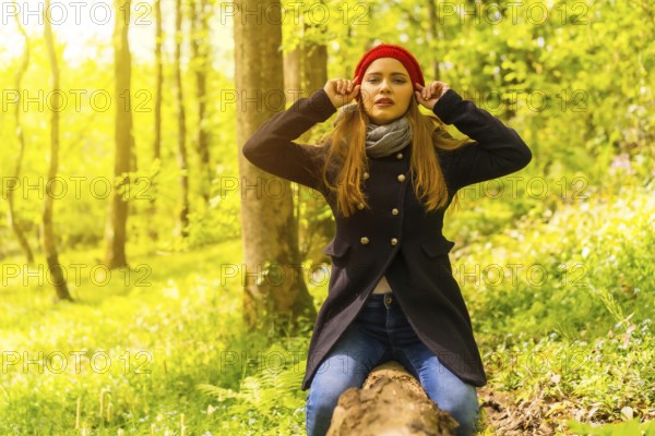 Beautiful woman with a black jacket and a red hat enjoying in an autumn park, sitting on a tree looking at camera