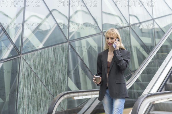 Entrepreneurial girl in business park in a modern building with escalator, young blonde in a suit making a call with a take away coffee