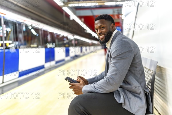 Young black businessman sitting on bench in subway station using smartphone while waiting for train