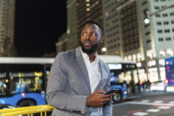 Young businessman holding a smartphone, looking around in a bustling city at night, surrounded by bright lights and modern skyscrapers