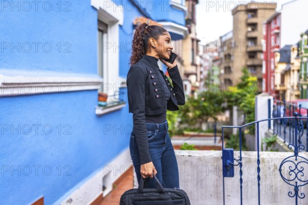 Side view photo of an african american casual businesswoman walking talking to the mobile among colorful street carrying a laptop bag