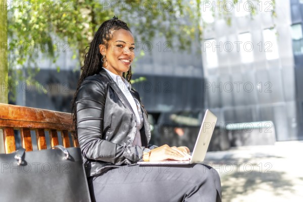Side view portrait of a successful latin businesswoman working with laptop sitting on city bench outdoors in a sunny day