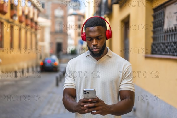 Frontal portrait of a serious young african man using mobile phone and headphones walking down street