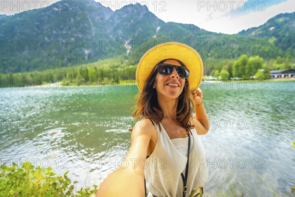 Tourist wearing sunglasses and a straw hat takes a selfie at lake dobbiaco with the dolomites in the background during a summer day