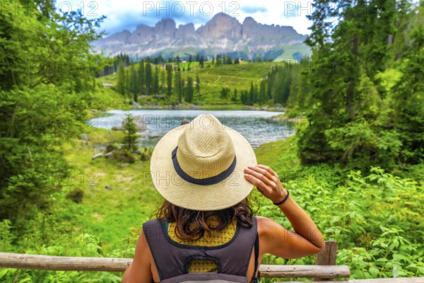 Female tourist wearing straw hat enjoying the landscape of lake carezza with latemar mountain range in the background in dolomites on a summer day