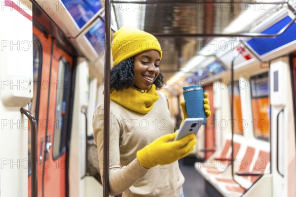 Smiling woman wearing yellow winter clothes using phone and holding reusable coffee cup while commuting on public transport
