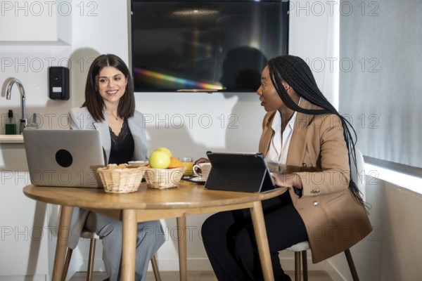 Two businesswomen are enjoying breakfast and working with laptop and tablet in their hotel room during a business trip