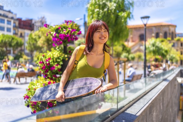 Smiling young chinese woman with skateboard gazing views form viewpoint in an urban square