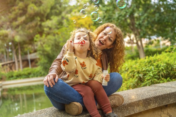 Caucasian happy mother and girl blowing soap bubbles sitting in the park