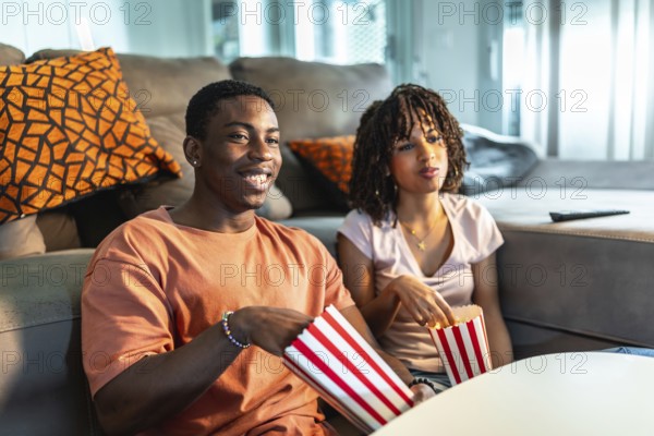 Young black couple enjoying a movie night at home, sitting on the sofa and eating popcorn from striped buckets