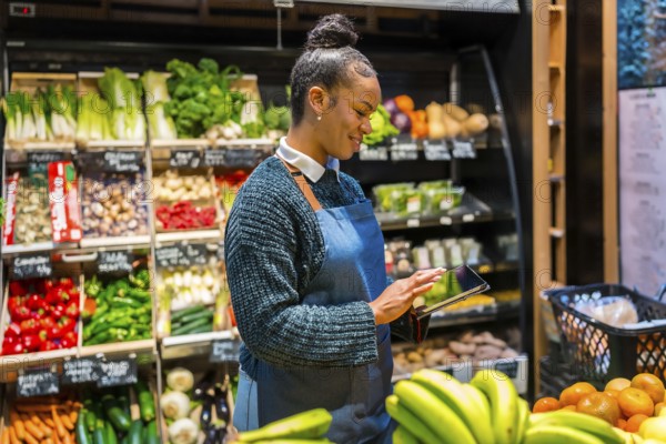 Smiling saleswoman wearing apron using digital tablet and managing inventory of fresh organic vegetables and fruits in supermarket