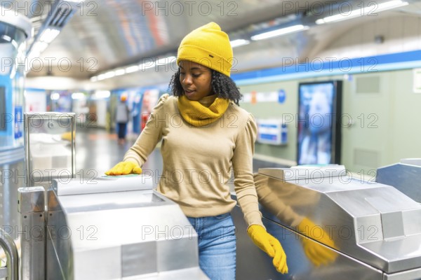 Young black woman wearing yellow woolen hat and gloves validating electronic ticket to enter subway station