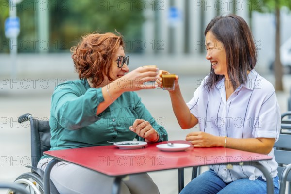 Woman with disability and asian friend toasting with coffee sitting on an outdoor cafeteria in the city