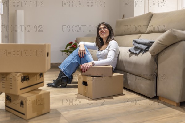 Young woman taking a break from unpacking, sitting on the floor of her new apartment and looking up dreamily