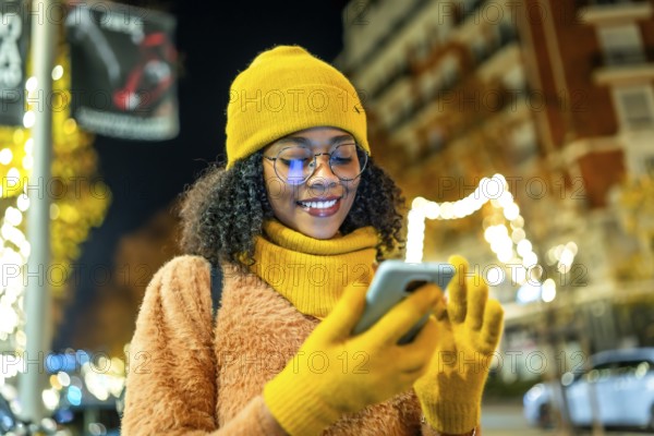 Smiling young woman wearing yellow winter clothes and using smartphone in a city street at night with christmas lights