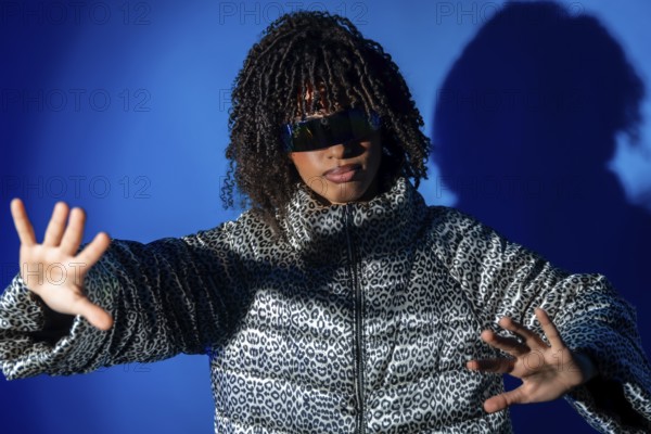 Fashionable woman with curly hair wearing futuristic sunglasses and animal print jacket making stop gesture with hands on vibrant blue background