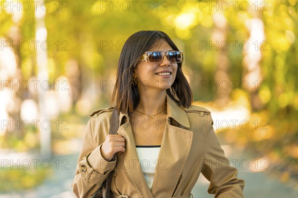 A woman wearing sunglasses and a brown coat is walking down a path. She is smiling and holding a handbag. Autumn or winter concept in the city