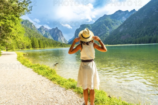 Tourist holding her straw hat admiring the stunning landscape of lake dobbiaco and the dolomites mountains in summer, italy