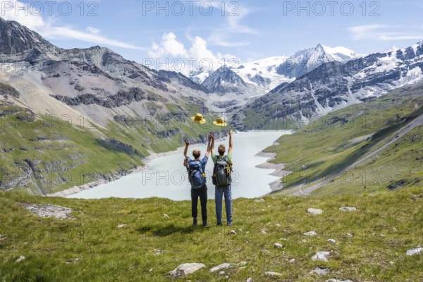 Two friends throwing yellow hats in the air, mountaineers in front of a blue mountain lake, Lac des Dix reservoir, behind mountain peak Mont Blanc de Cheilon with glacier, Hérménence, Valais Alps, Valais, Switzerland, Europe