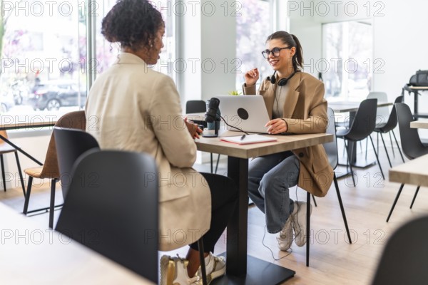 Two women engaging in a podcast recording session at a cafe, utilizing a laptop and microphone for their lively discussion