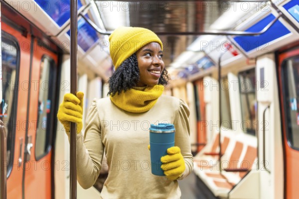 Smiling young woman wearing yellow winter clothes and holding reusable coffee cup standing inside subway train