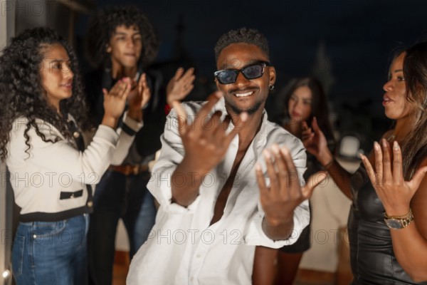 Brunette cool man in sunglasses gesturing while dancing surrounded by friends at night in a city roof