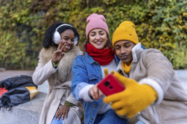 Three happy young students are taking a selfie in a city park during winter, wearing warm colorful clothing