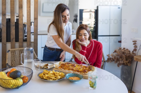 Two women enjoying a meal together, sharing pizza, tacos, nachos, and other snacks in a bright, modern kitchen