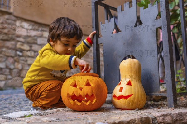 A young boy is playing with two pumpkins, one of which has a scary face. The scene is set in a courtyard, with a stone wall in the background. The boy is wearing a yellow shirt and brown pants